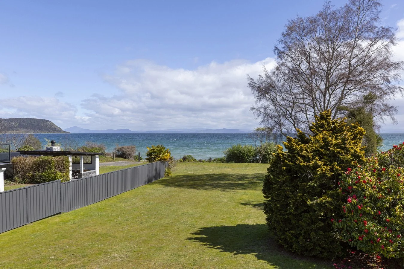A backyard with green grass, bushes, and trees, overlooking an ocean with distant islands and a partly cloudy sky.