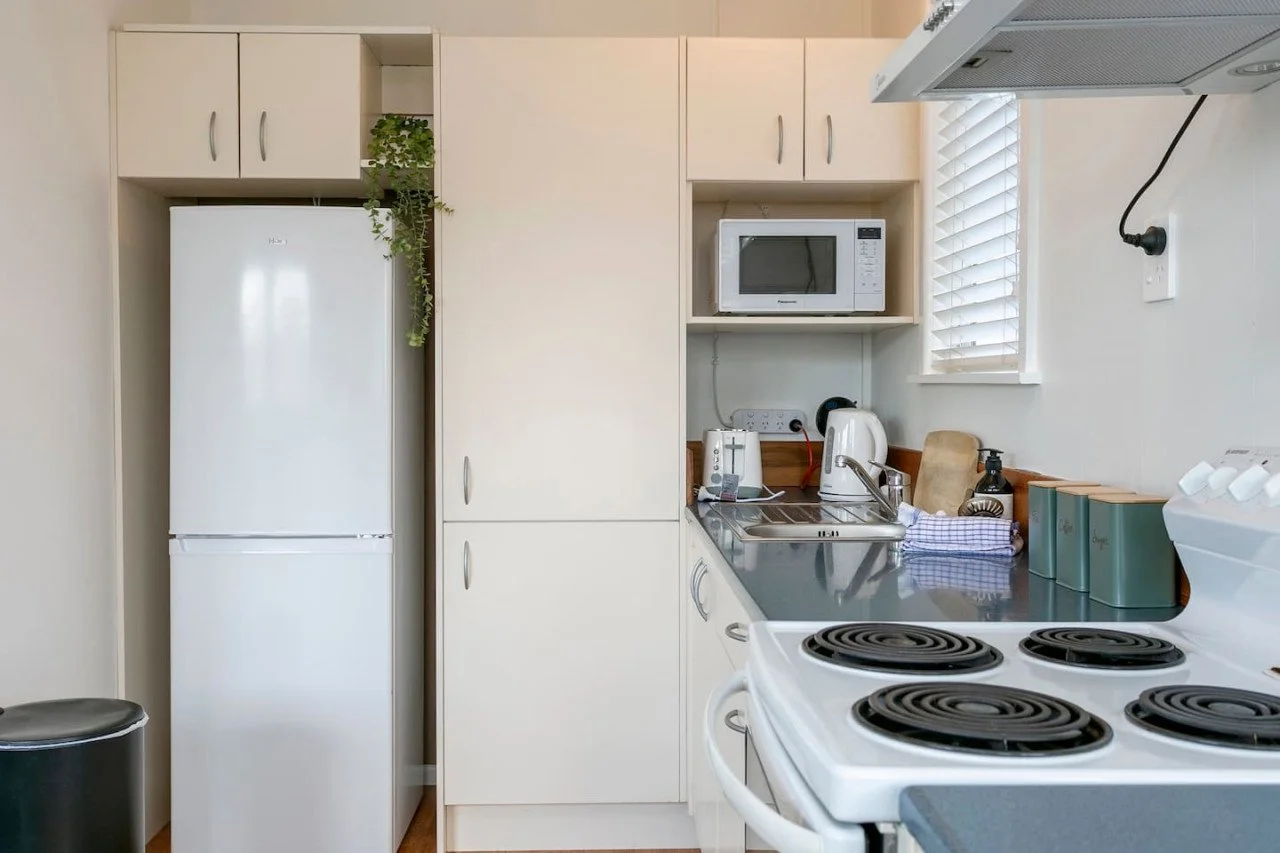 Compact kitchen with white appliances, including a refrigerator, microwave, and stove. Countertop holds a tea kettle, soap dispenser, and a dish towel. There are cabinets, an electric outlet, and a window with blinds.