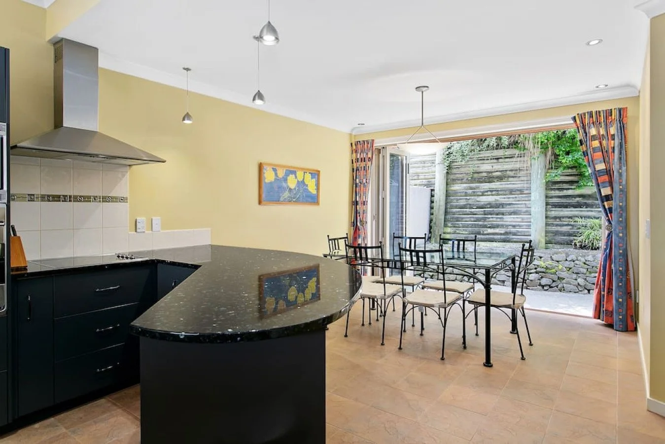 Open kitchen and dining area with a black countertop bar, yellow walls, and a sliding glass door leading to an outdoor garden with a wooden fence and rocks.