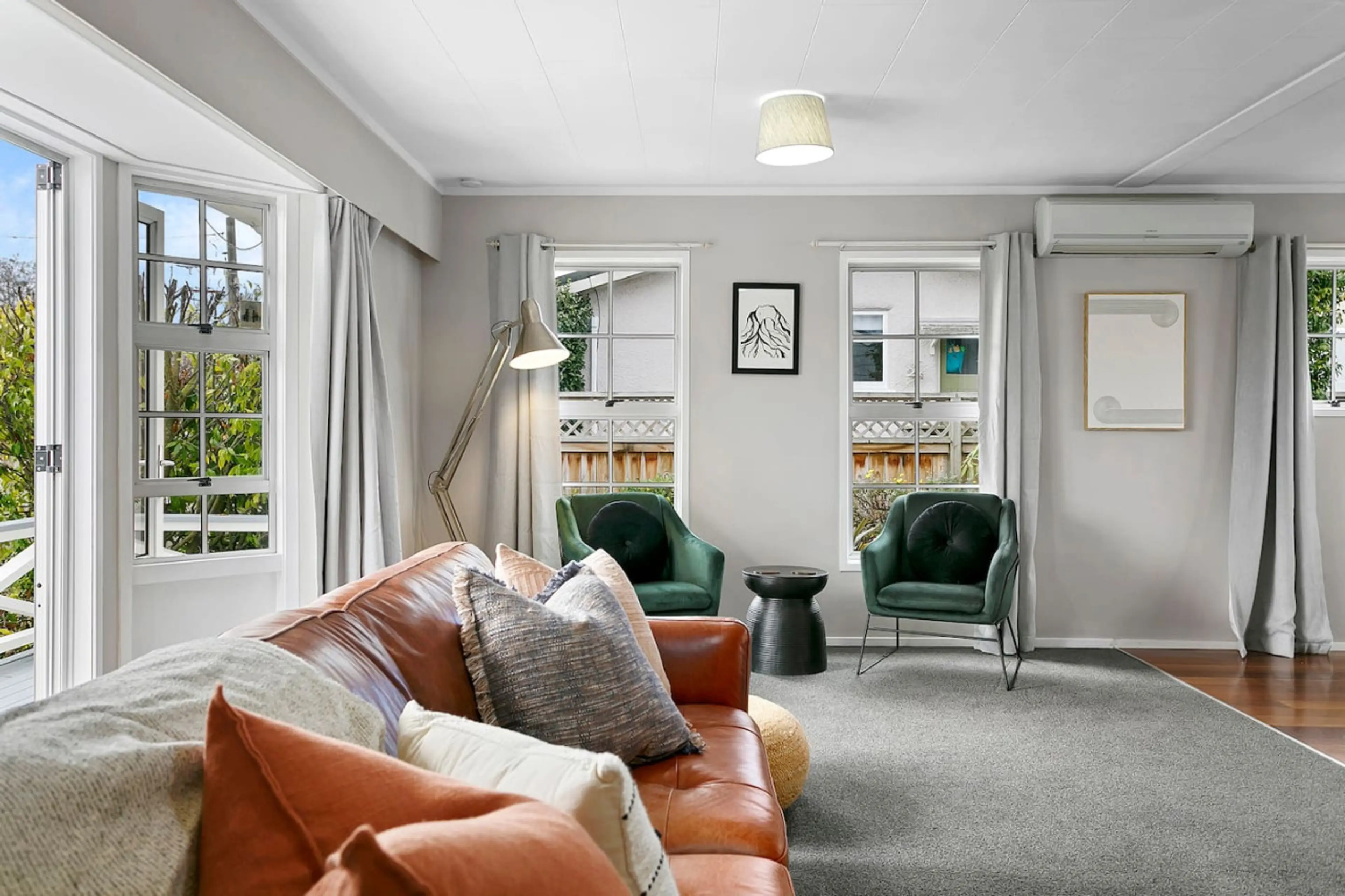 Living room with beige sofa, green armchairs, side table, large windows with white curtains, and wall art.
