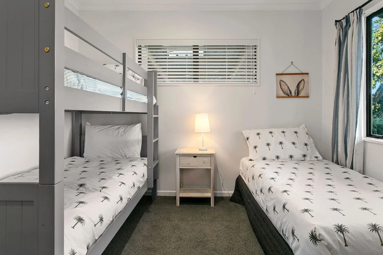 Bedroom with a gray bunk bed and a white bed, both with palm tree patterned bedding, a nightstand with a lamp, window with blinds, and a second window with curtains.