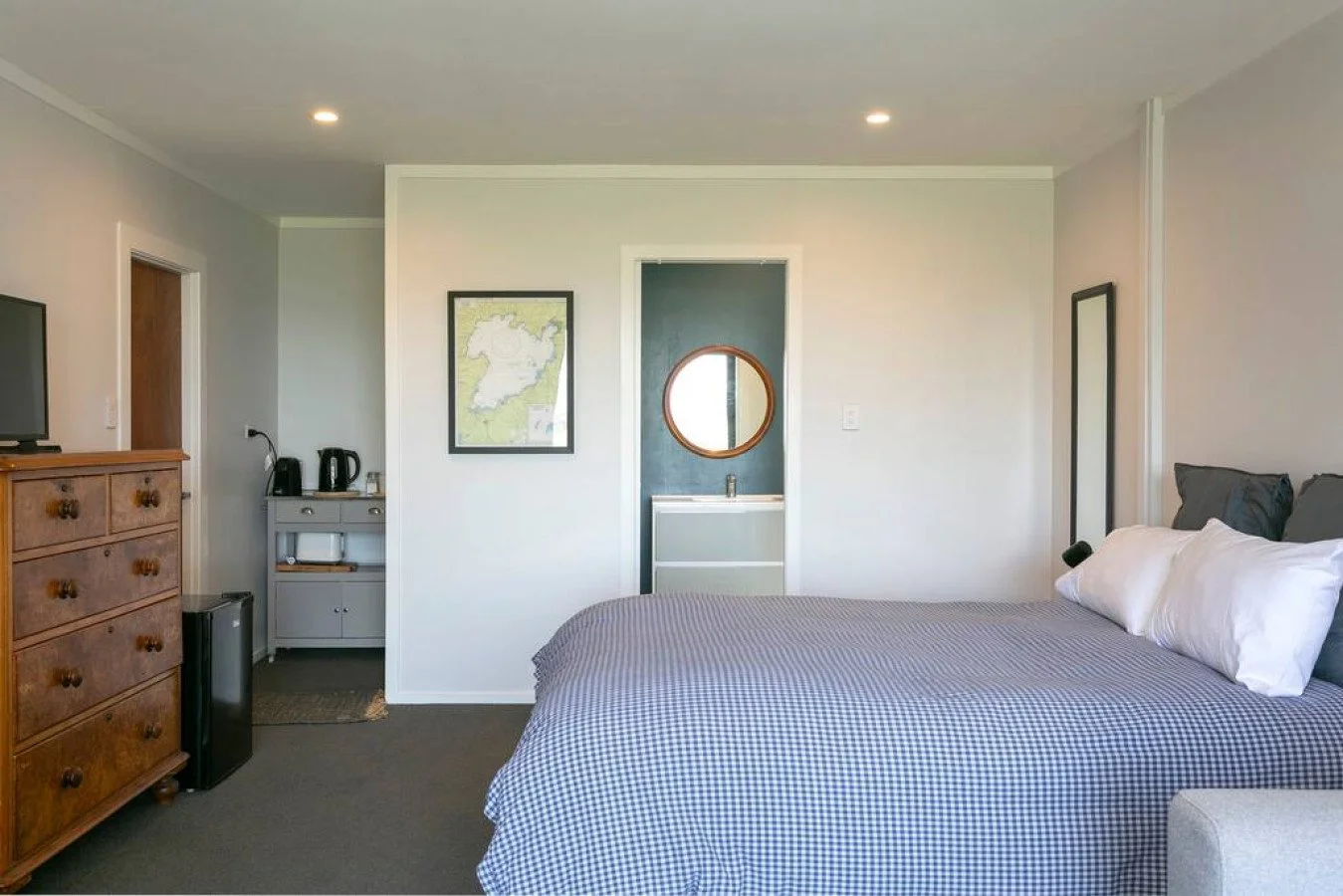 A bedroom with a bed, pillows, a dresser, a TV, and a small kitchenette area with a map and kettle, with a doorway leading to a small bathroom with a mirror.