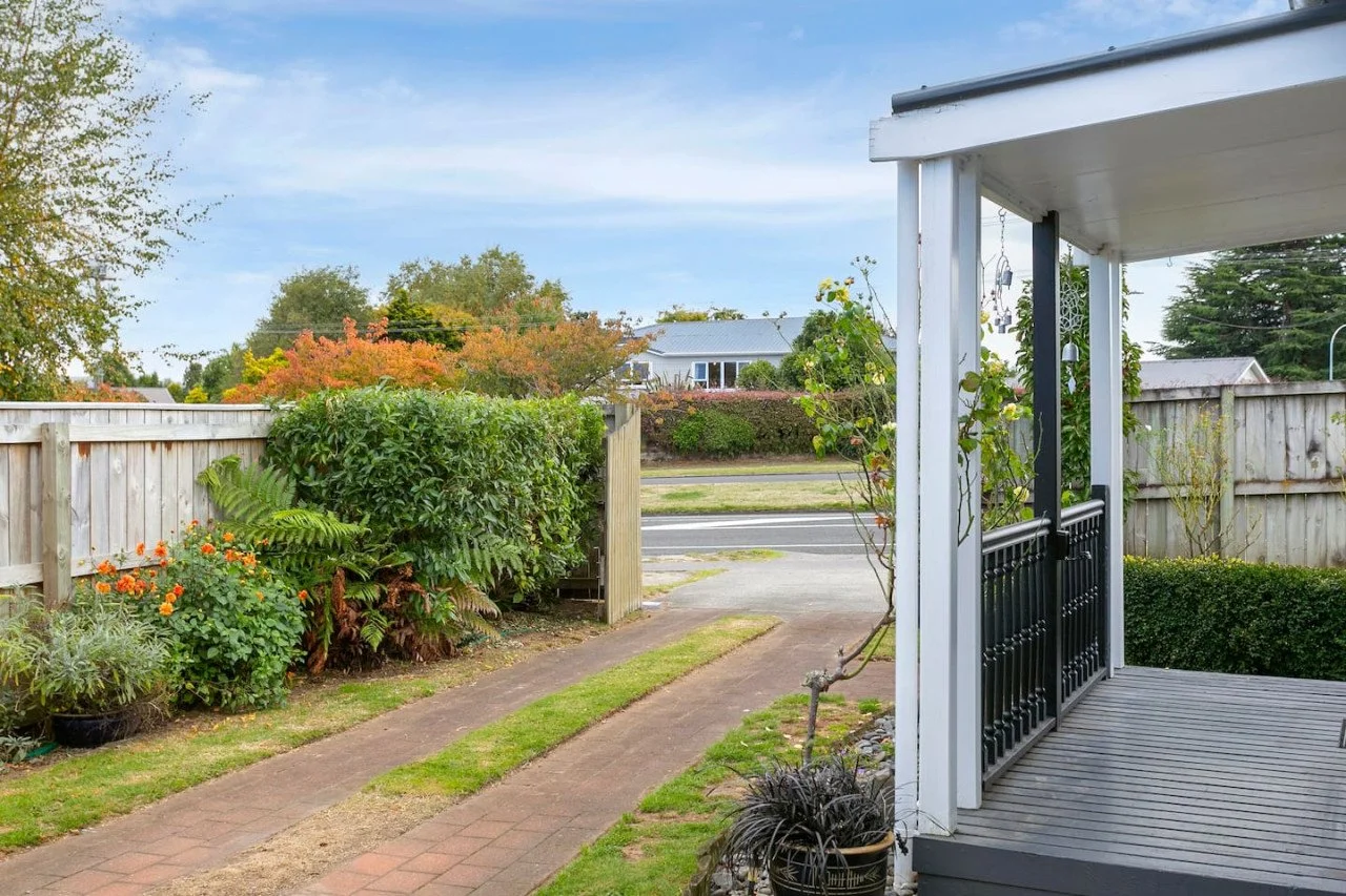 Front yard view showing a sidewalk leading to a wooden fence, bushes, plants, and a porch with a railing, with a street and houses in the background under a partly cloudy sky.
