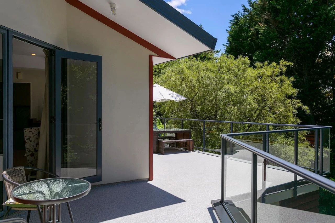 A spacious outdoor balcony attached to a house, with a glass table and chairs, overlooking trees and greenery, with an umbrella and outdoor seating in the background.