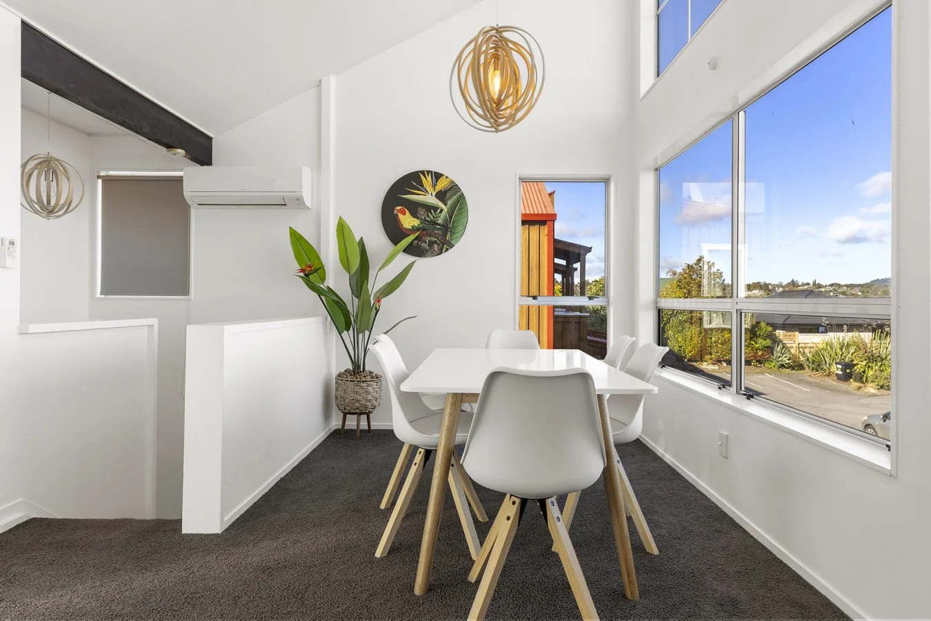 Bright dining area with white chairs, a white table, large windows showing an outdoor view, a potted plant in a wicker basket, and modern light fixtures.