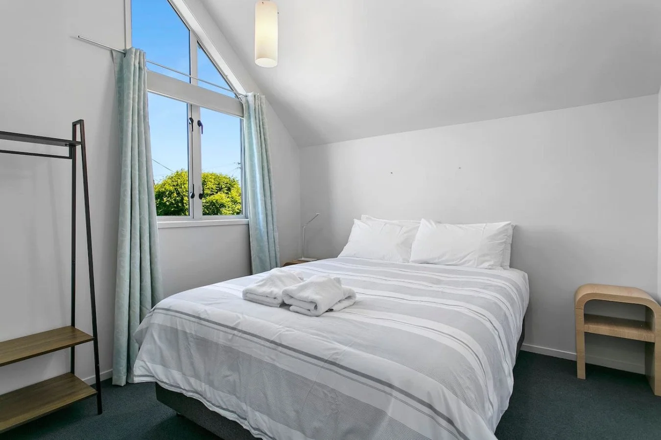 Clean hotel bedroom with white bedding, pillows, and towels on the bed, window showing outdoor trees, and minimal furniture.