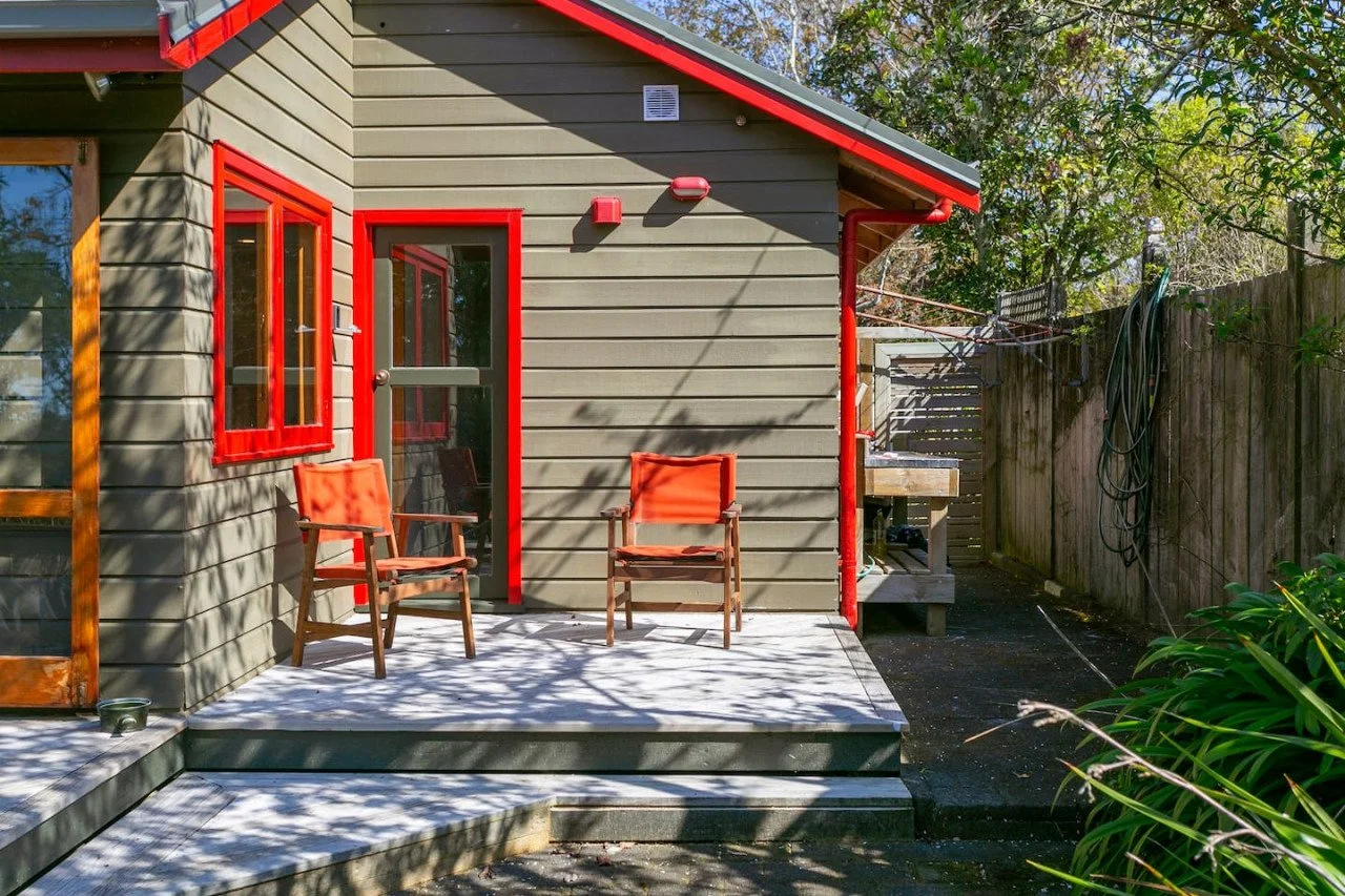 Backyard patio of a house with two wooden chairs with orange cushions on a small concrete porch, a door and window with red frames, and a wooden fence with garden hose and greenery.
