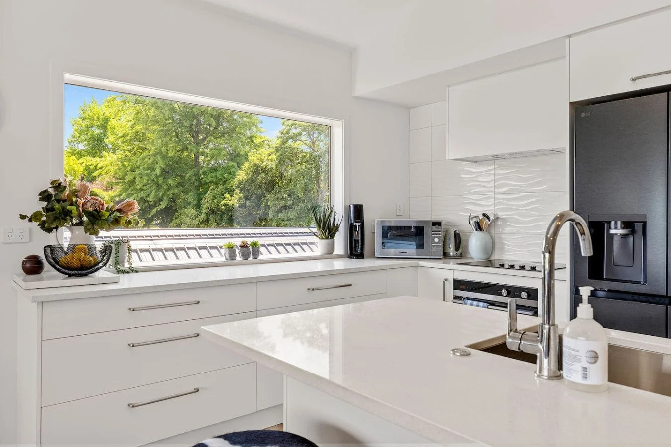 Modern white kitchen with large window showing green trees outside, with appliances and decor on the counter.