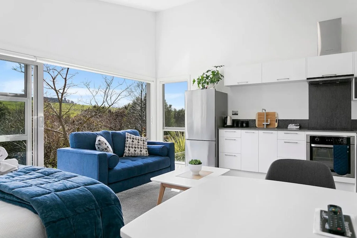 Modern living space with large windows, a blue sofa, a bed, a white table with a remote, and a white kitchen with black accents.