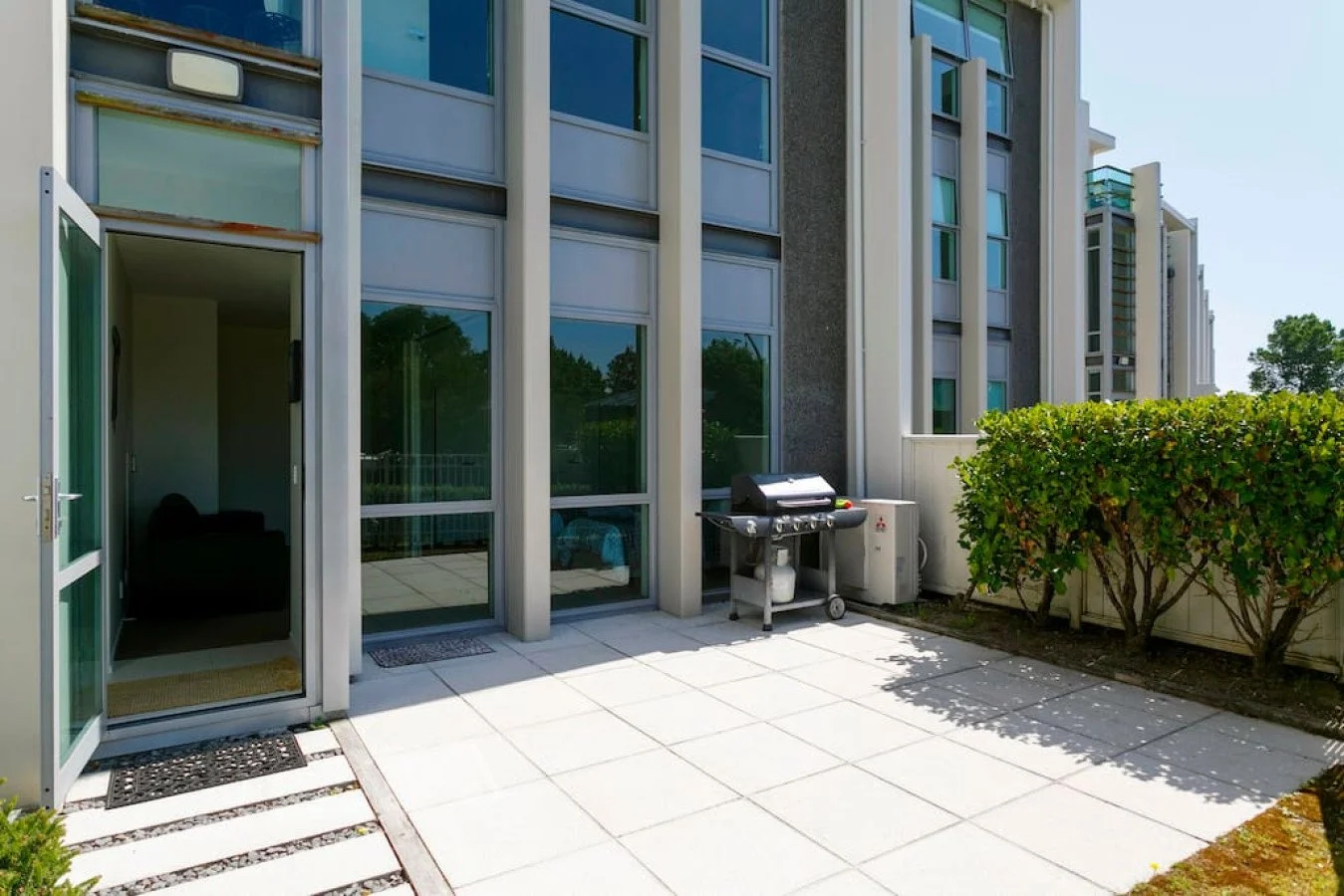 Outdoor patio area of an apartment building with a barbecue grill, a small hedge, and glass sliding doors.