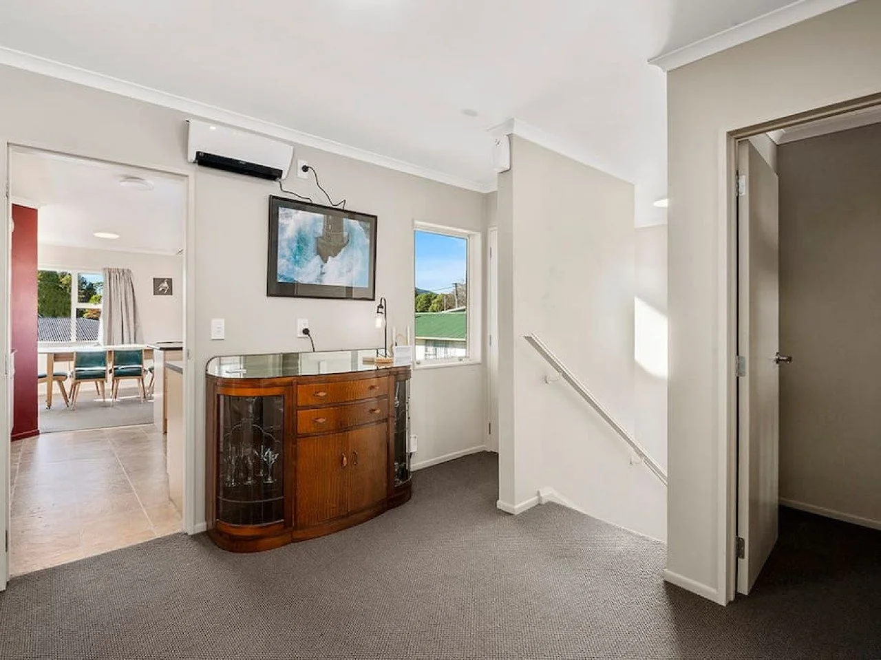 Interior view of a house showing a hallway with a vintage wooden sideboard, framed artwork on the wall, a window with a view of neighboring houses, and a staircase leading down.