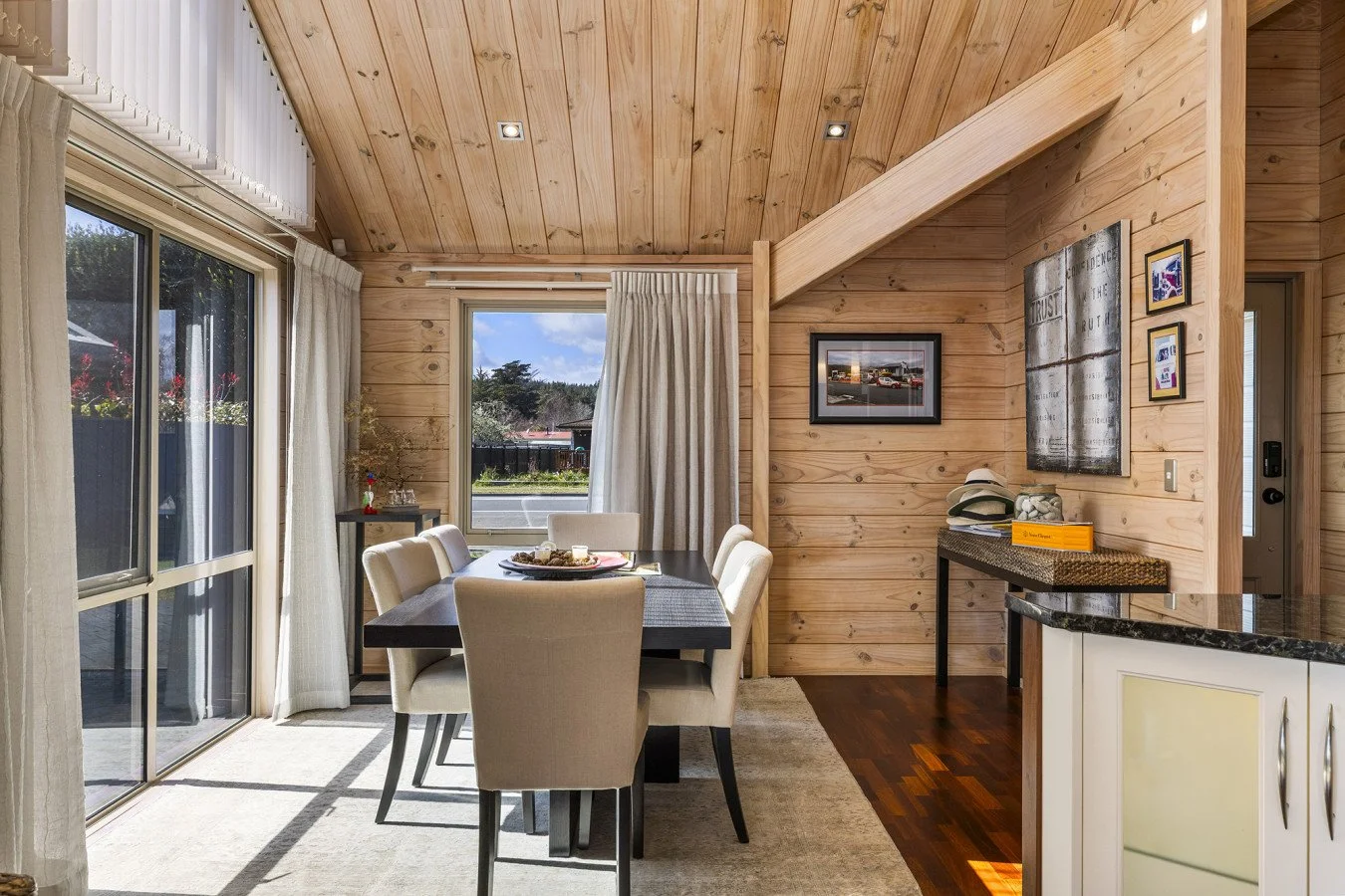 Dining area with a dark wood table and six beige upholstered chairs, near large sliding glass doors and windows with white curtains, and wood-paneled walls and ceiling, decorated with framed pictures and artwork.