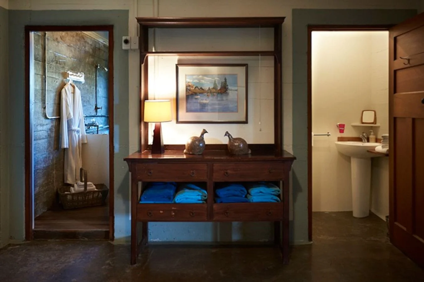 Interior view of a room with a wooden dresser topped with two duck sculptures, a framed landscape painting, and a lamp. To the left, a bathroom with a hanging robe, a sink, and a mirror. To the right, another bathroom with a sink and toiletries.