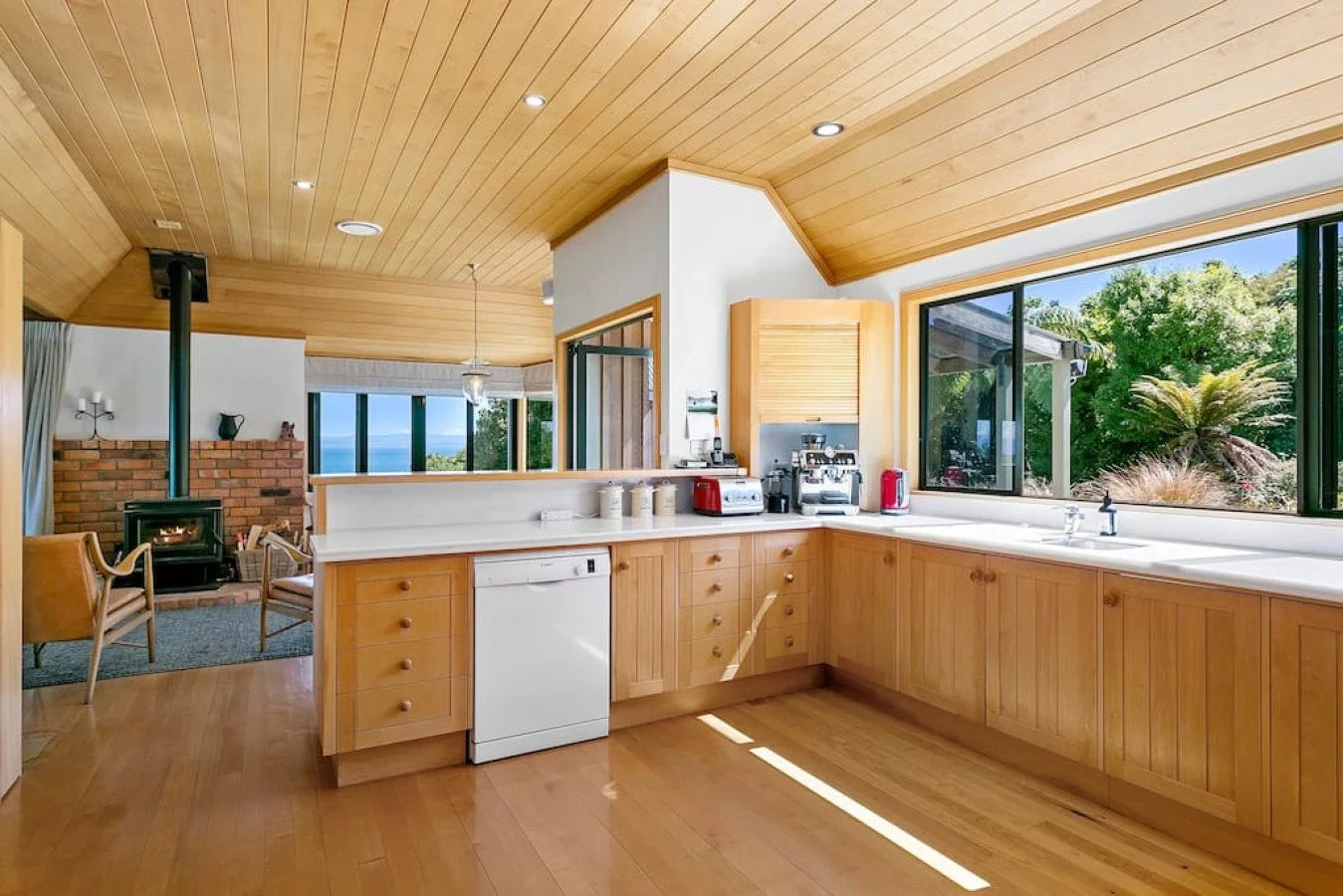 Kitchen with wooden cabinets, large window showing outdoor greenery, and a view into a living room with a brick fireplace and chairs.