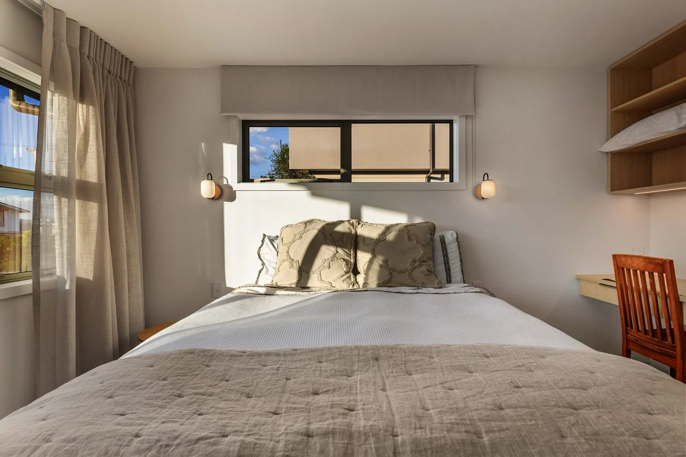 A bedroom with a bed, pillows, and beige bedding, illuminated by natural sunlight through a window, with wall-mounted lamps and a wooden desk and chair.