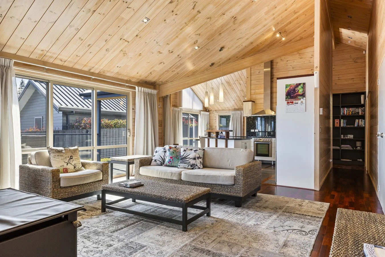Living room with large windows, wooden ceiling, and kitchen in the background. Contains a sofa, armchair, decorated pillows, coffee table, and a patterned rug.