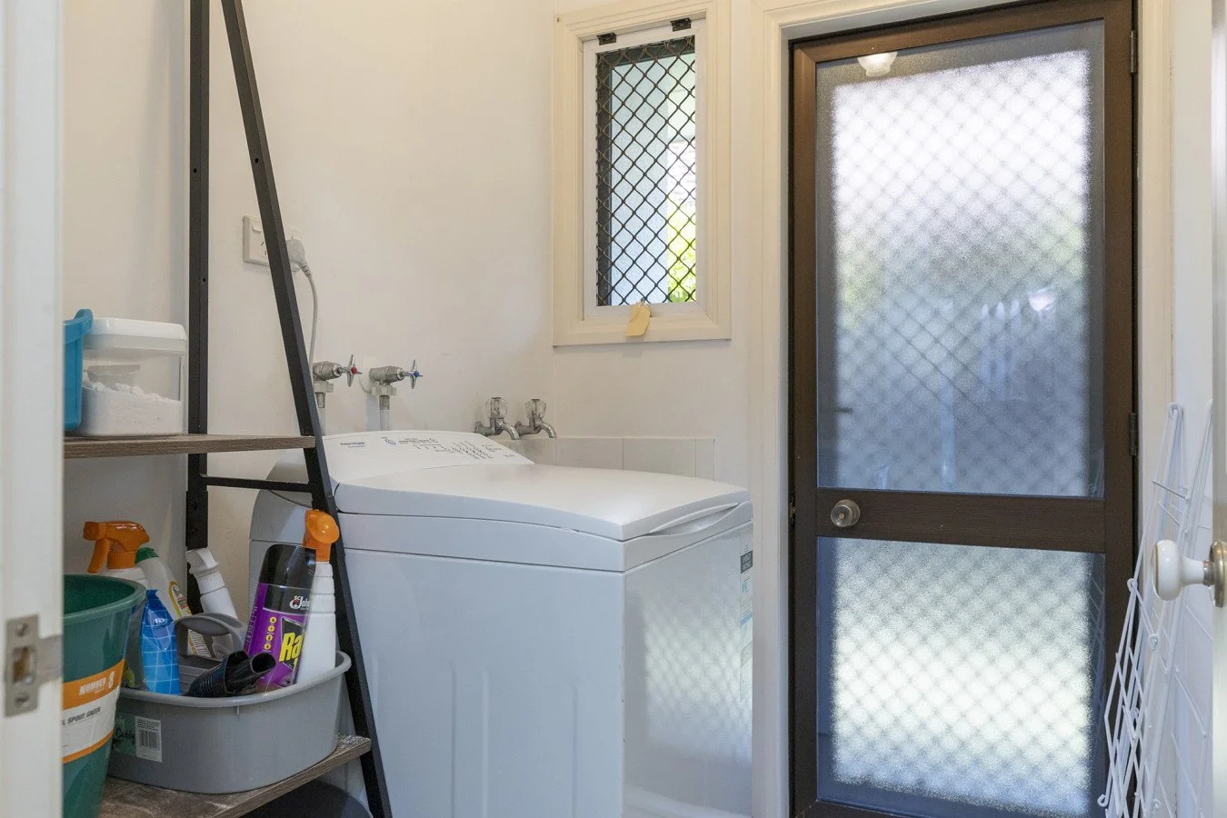 Laundry room with a top-loading washing machine, a small window with security bars, a glass door with a security screen, and shelves with cleaning supplies.