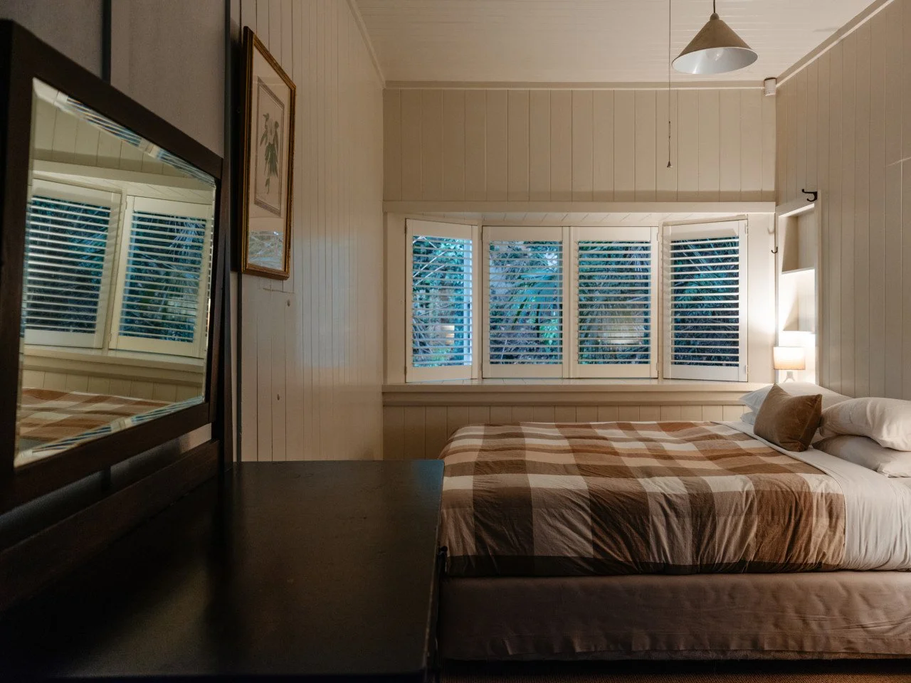 A cozy bedroom with white wooden panel walls, a bed with a checkered bedspread, a side table with a lamp, a window with white shutters, and a mirror reflecting part of the room.