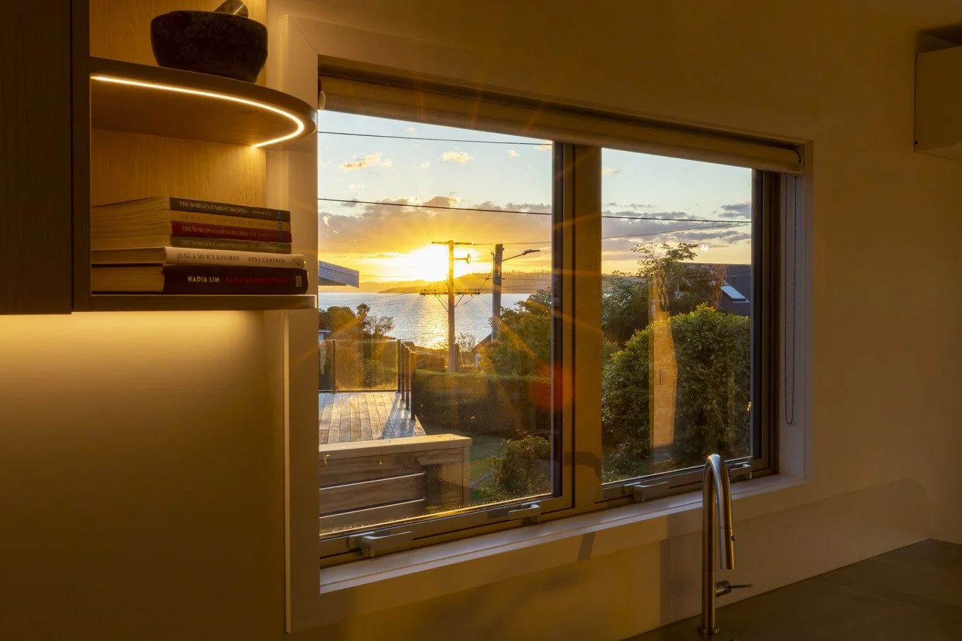 Sunset view through a kitchen window, with houses and greenery outside, and a shelf with books on the left.