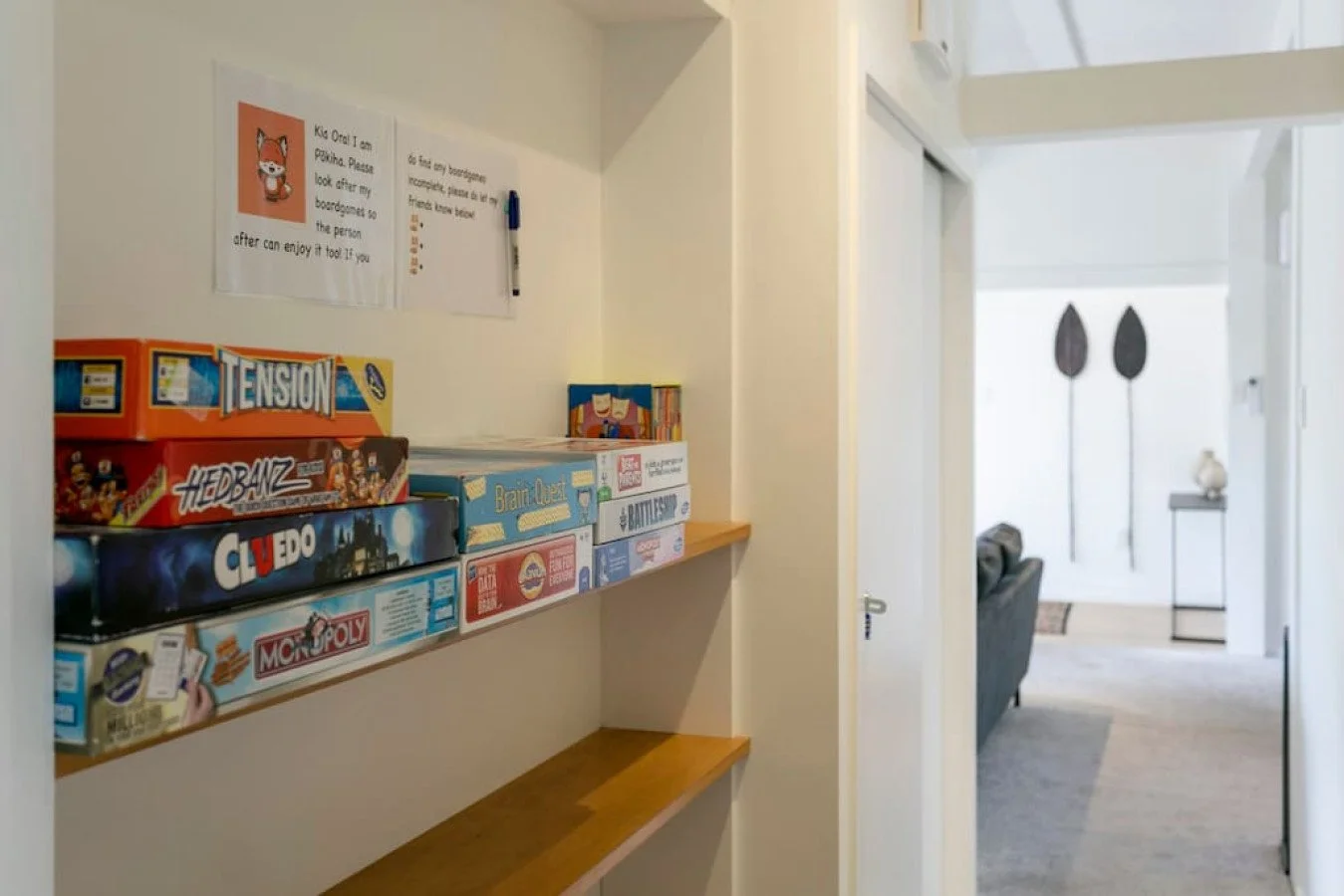 Board game shelf in a hallway with various board games including Monopoly, Cluedo, Hedbanz, and Tension, with a notice on the wall above the shelf.