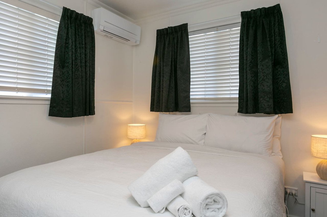 A neatly made bed with white linens and pillows, with rolled towels placed at the foot of the bed. The room has three windows with white blinds and black curtains, and two bedside lamps providing warm lighting.
