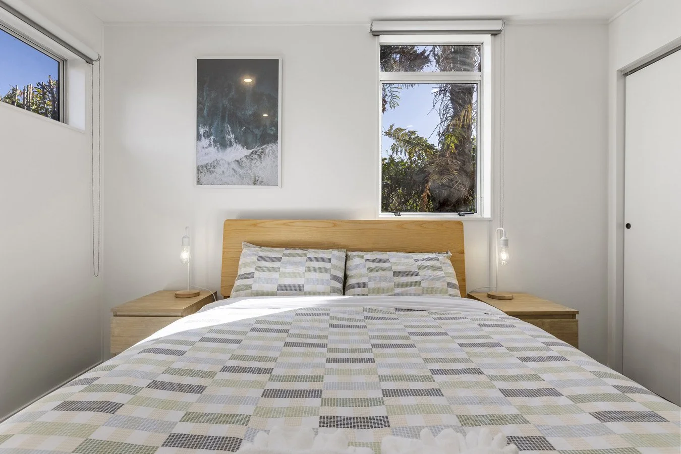 Modern bedroom with white walls, wooden bed frame, and checkered bedding. Two bedside tables with hanging lamps. Window showing trees outside, framed artwork on wall.