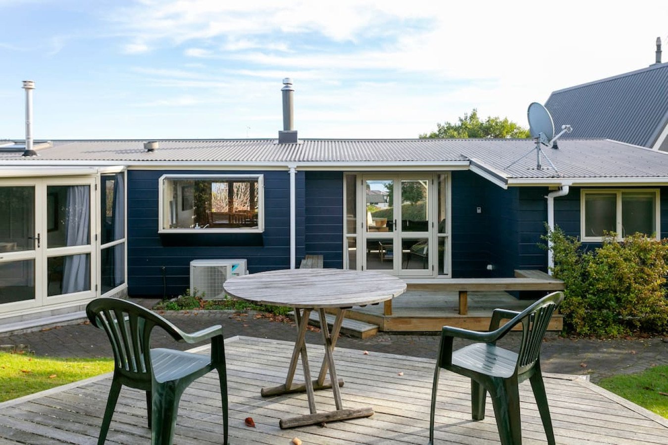 Backyard patio with a wooden table and two green chairs, adjacent to a blue house with sliding glass doors and windows, and a satellite dish on the roof, under a blue sky with some clouds.