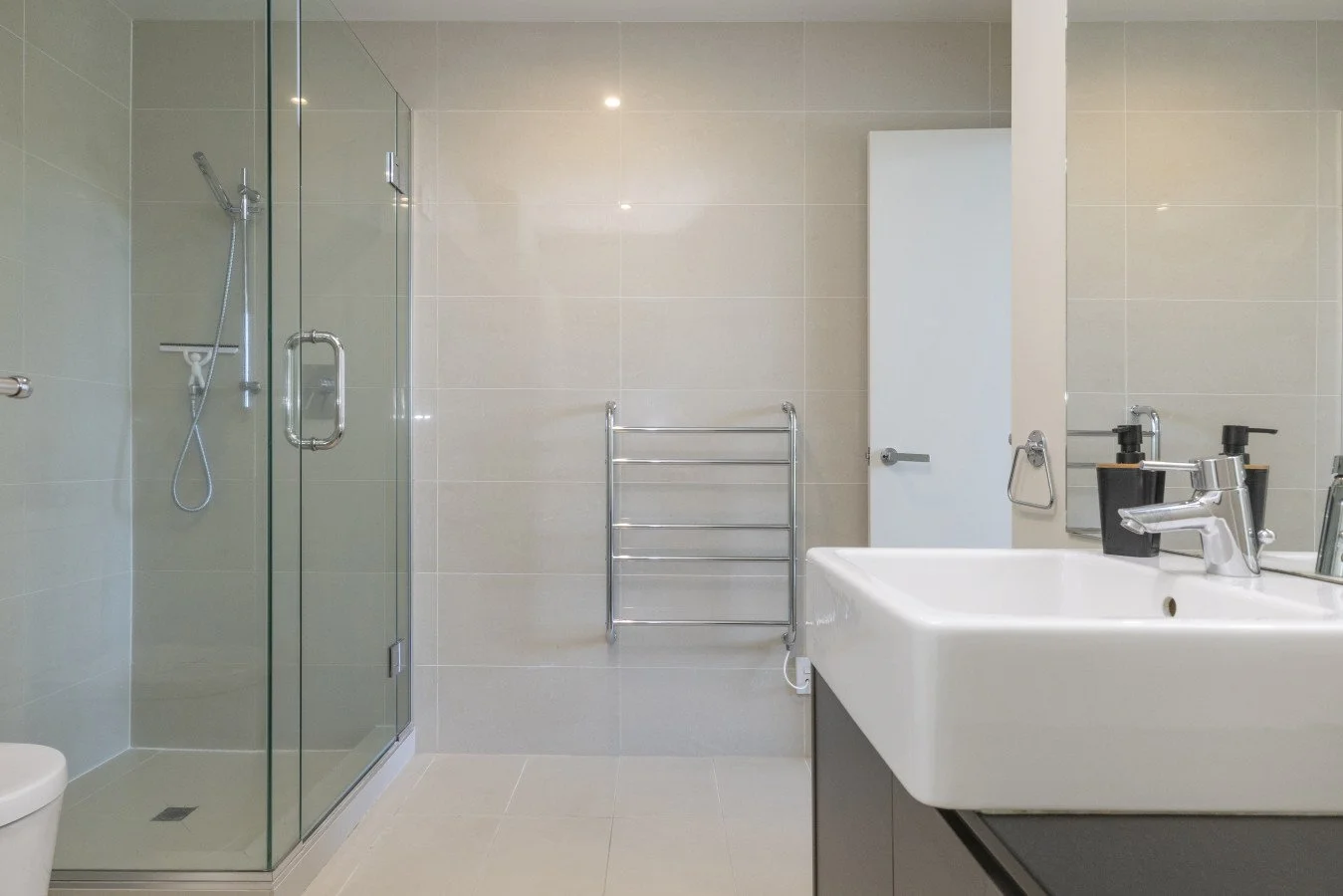 Modern bathroom with a glass-enclosed shower, white toilet, white sink with a chrome faucet, black soap dispensers, a wall mirror, and a stainless-steel towel rack.