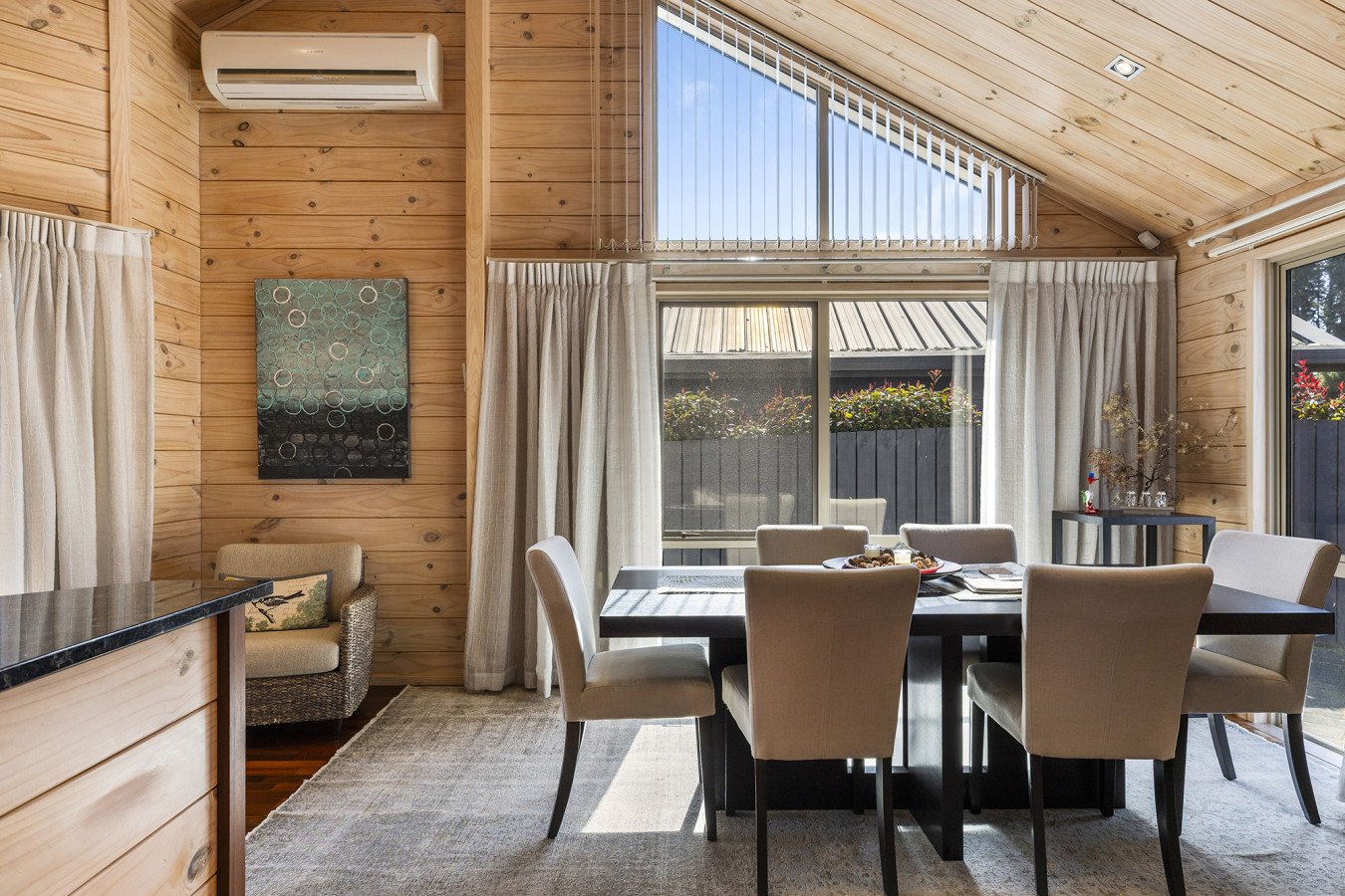 A dining area with beige upholstered chairs around a black table, a large window with white curtains, a wooden wall with a painting, and an air conditioning unit in a room with wooden paneling.