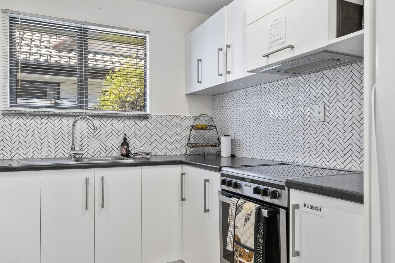 Kitchen with white cabinets, black countertop, herringbone tile backsplash, window with blinds, stove with towels hanging, small rack with fruit, paper towel roll, and a 