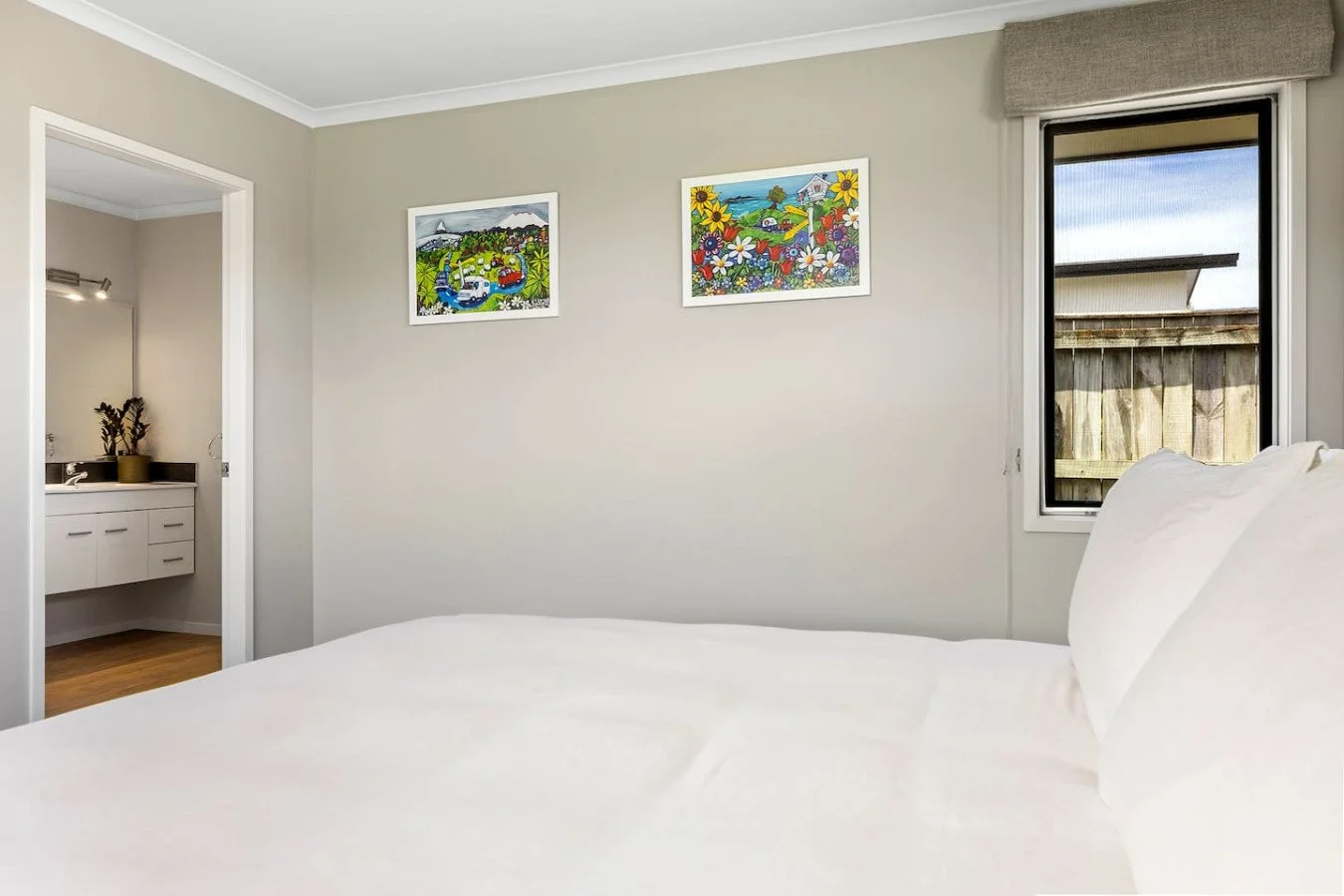 A bedroom with white bedding, a window with a view of a wooden fence, and colorful framed pictures of landscapes and flowers on the wall.