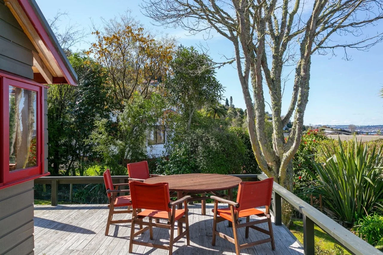 A wooden deck with a round table and four red chairs, surrounded by trees and plants, with a view of a distant landscape on a clear day.