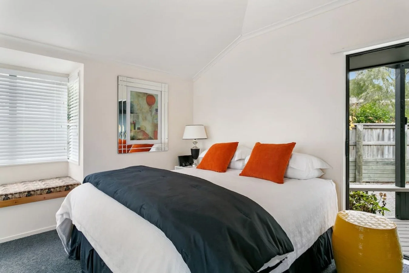 A bedroom with a large bed featuring white sheets, two orange pillows, and a black blanket, positioned against a white wall with a mirror, artwork, and a white lamp on a side table. The room has a window with white blinds, a yellow textured ottoman, 