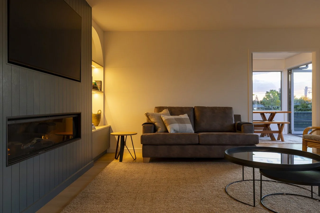 Living room with a dark gray sofa, a round black coffee table, a built-in fireplace, a flat-screen TV, a small round side table, and a shelf with decor items illuminated by soft lighting; sliding glass doors lead to a balcony with a picnic table, wit