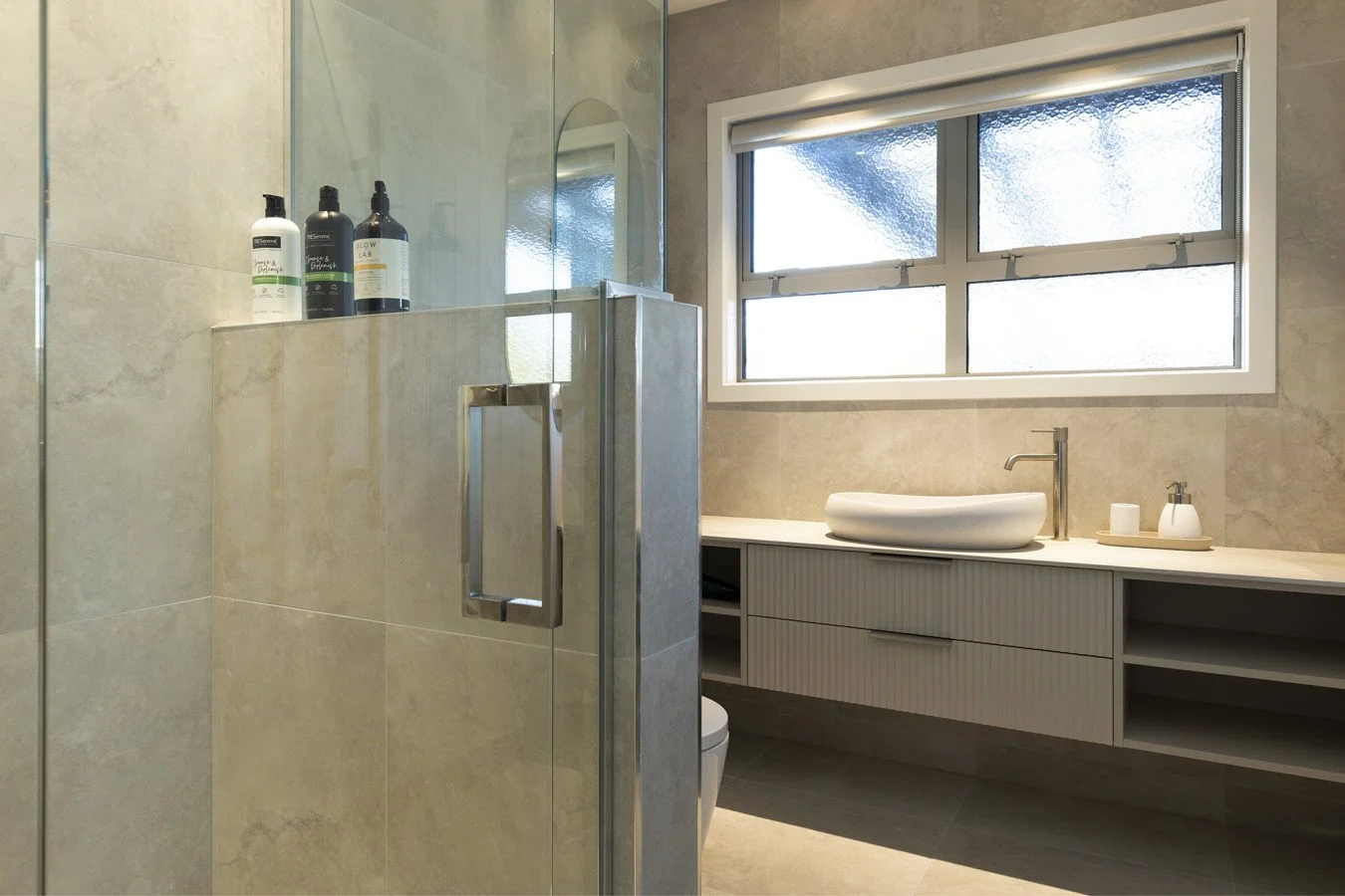Modern bathroom with a glass shower, large window, and a beige countertop with a vessel sink and a faucet.