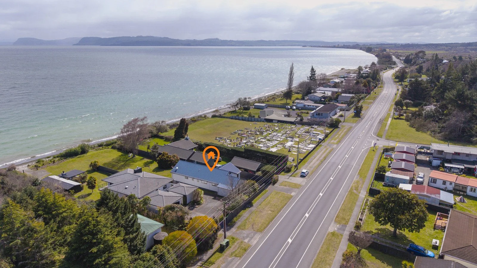 Aerial view of a coastal neighborhood with houses, a cemetery, and a road, next to the ocean under cloudy skies.