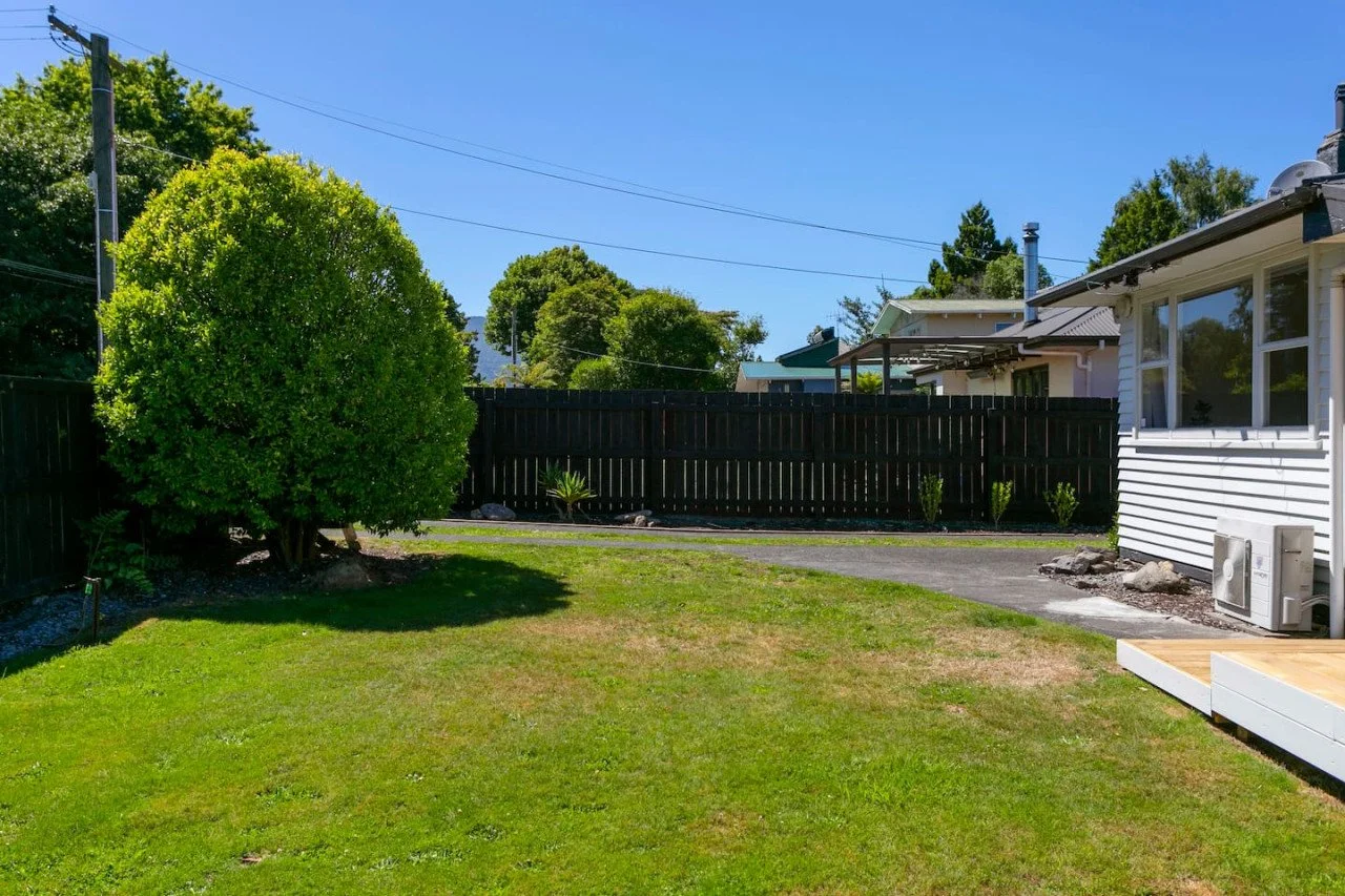 A backyard with a lush green lawn, a large bush on the left, a paved pathway, a black wooden fence, and part of a white house with windows and a small deck on the right, under a clear blue sky.