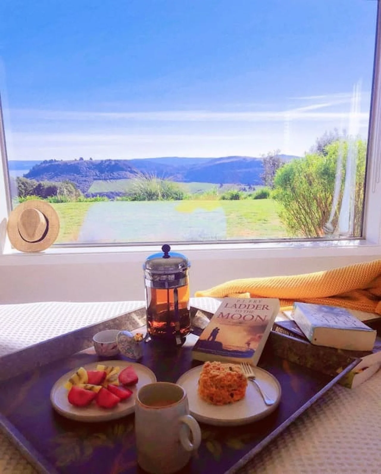 A breakfast tray with cookies, bread, and a cup of tea or coffee, set on a window sill with a scenic view of green hills and blue sky.
