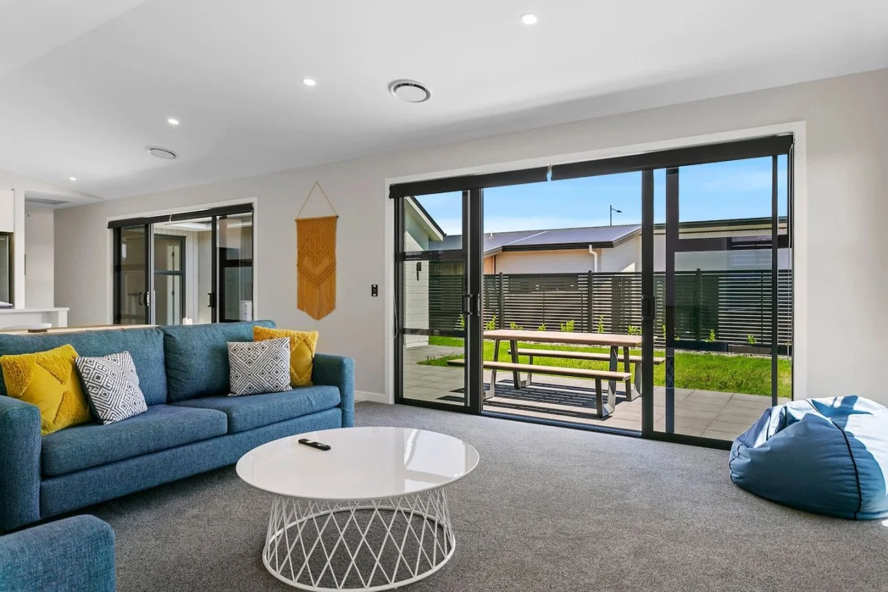 Living room with a blue sofa adorned with yellow and patterned pillows, a white coffee table, and a bean bag chair, with large sliding glass doors leading to an outdoor patio and backyard.
