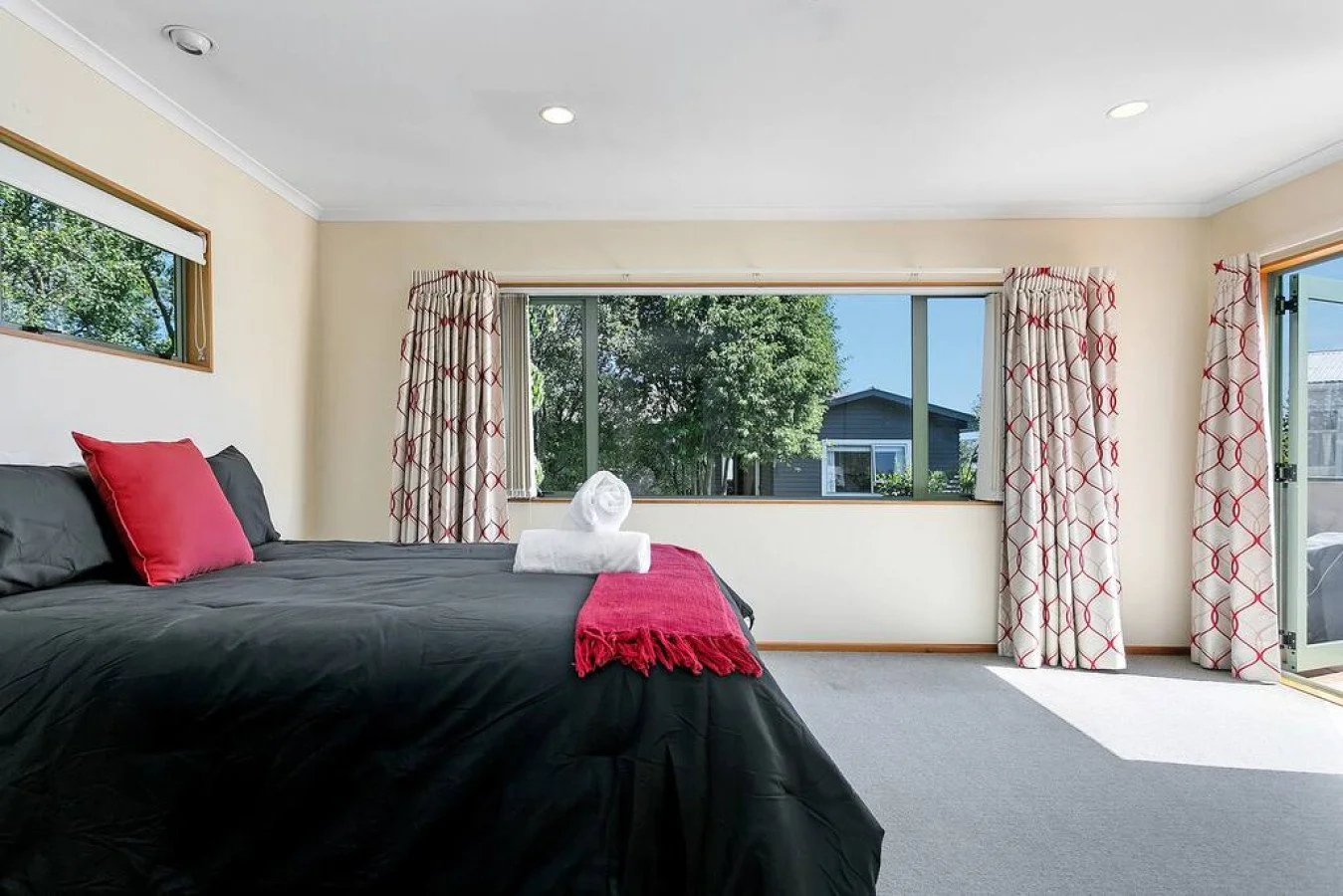A bedroom with a large window, a bed with black bedding and red pillows, and curtains with a red pattern. The room has beige walls, gray carpet, and a view of trees and neighboring houses outside.