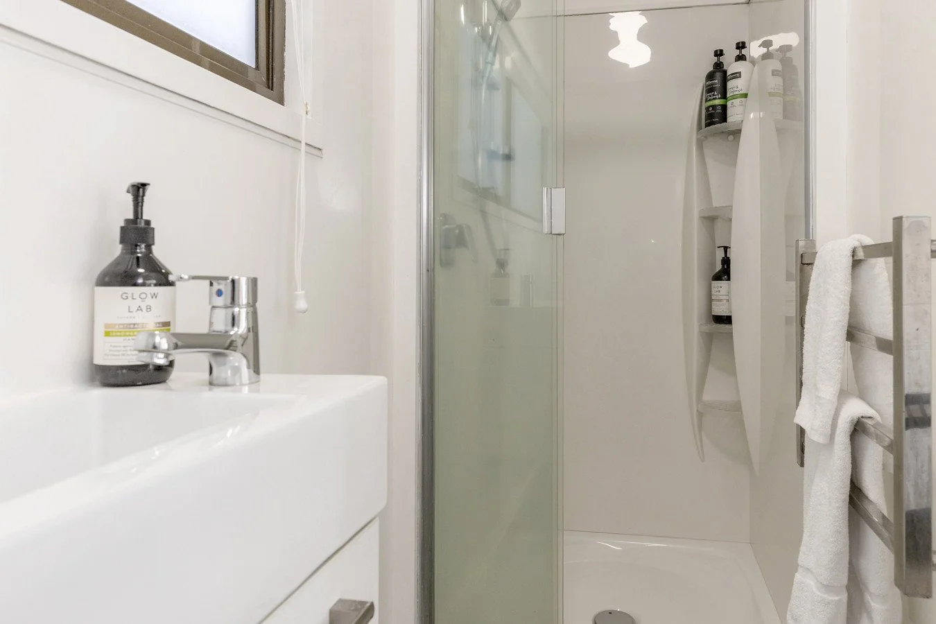 A small bathroom with a white sink, a silver faucet, a soap dispenser, a shower with a glass door, and built-in shelves holding bottles of soap or shampoo, along with towels hanging on a metal towel rack.