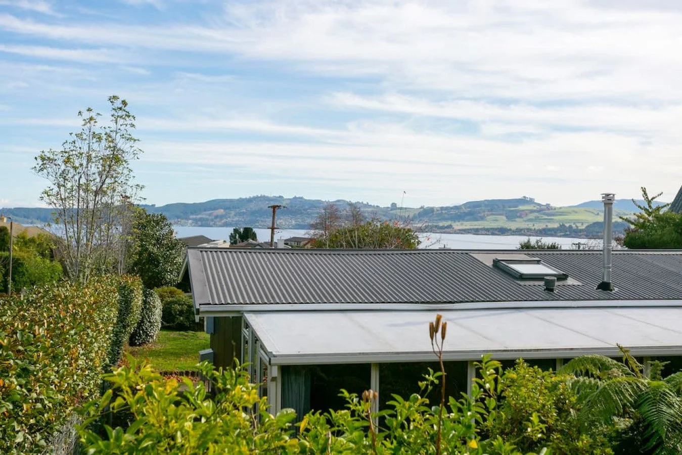 View of rooftops, trees, and a body of water with hills in the background under a partly cloudy sky.
