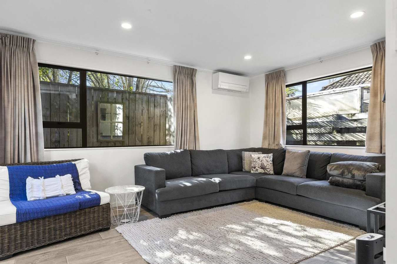 Living room with large gray sectional sofa, white and wicker armchair with blue throw, small white side table, beige curtains, windows showing outside wooden fence and garden, rug, and ceiling lights.