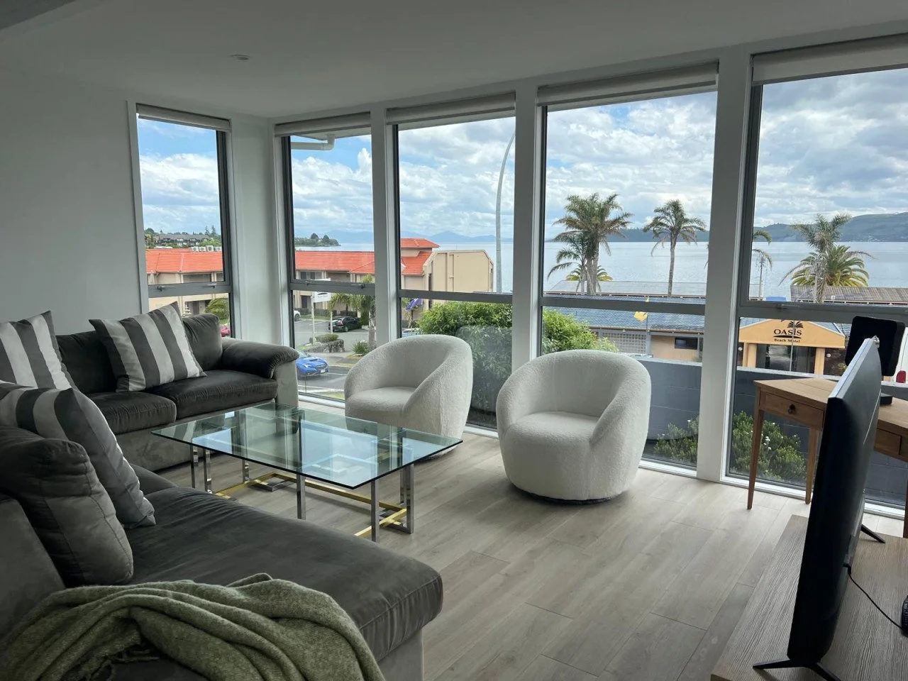 Living room with large windows overlooking water, featuring a gray sofa with striped pillows, two white rounded chairs, a glass coffee table, and a view of palm trees and buildings outside.