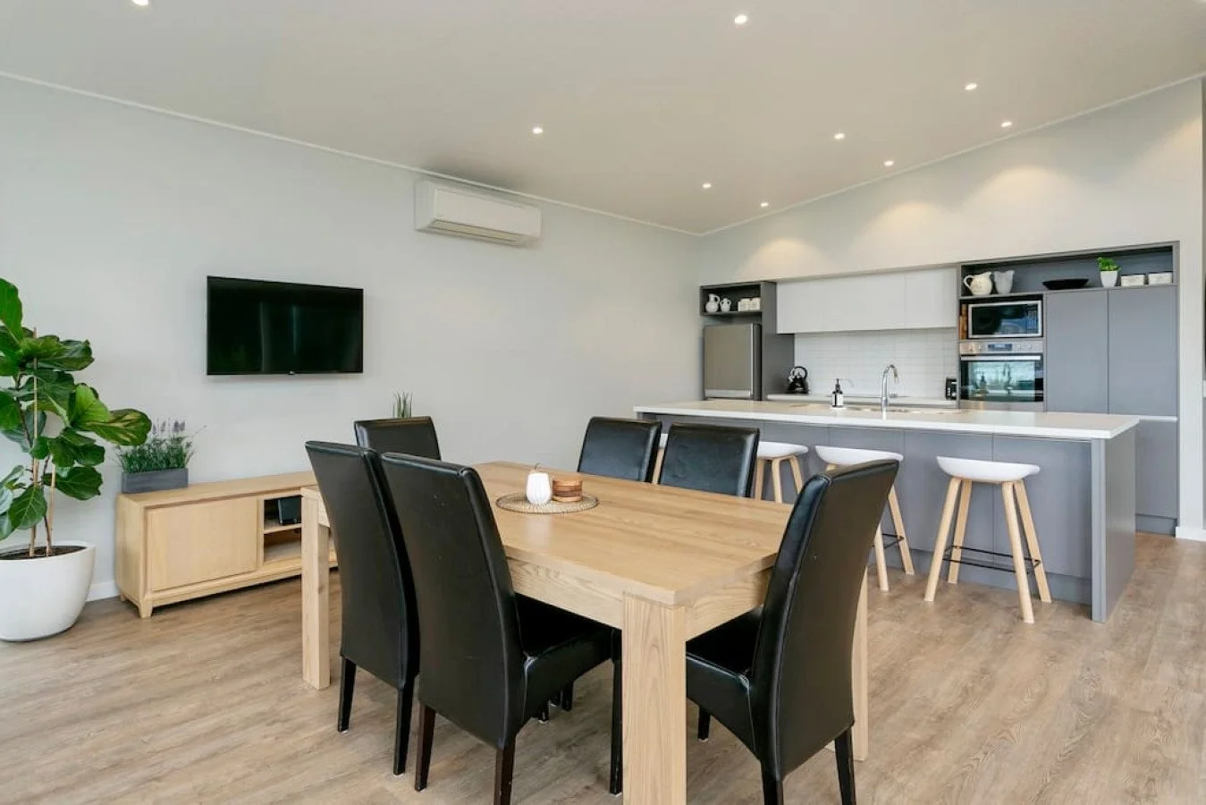 Modern dining room and kitchen with wooden floor, black chairs, a large wooden dining table, a wall-mounted TV, a kitchen island with white stools, and cabinetry with built-in appliances.