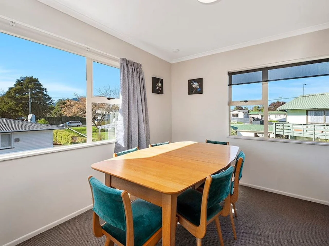 Dining room with wooden table, teal chairs, large windows showing neighborhood and blue sky, with curtains and wall art.