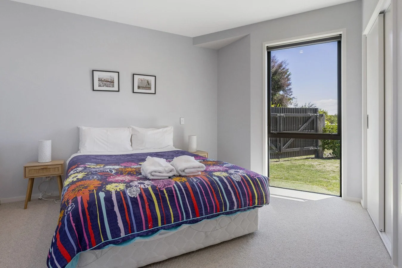 A bedroom with a bed covered in a colorful striped quilt, two white pillows, two folded white towels on the bed, two small bedside tables with white lamps, a large window with a view of a fenced backyard, and two framed pictures on the wall above the