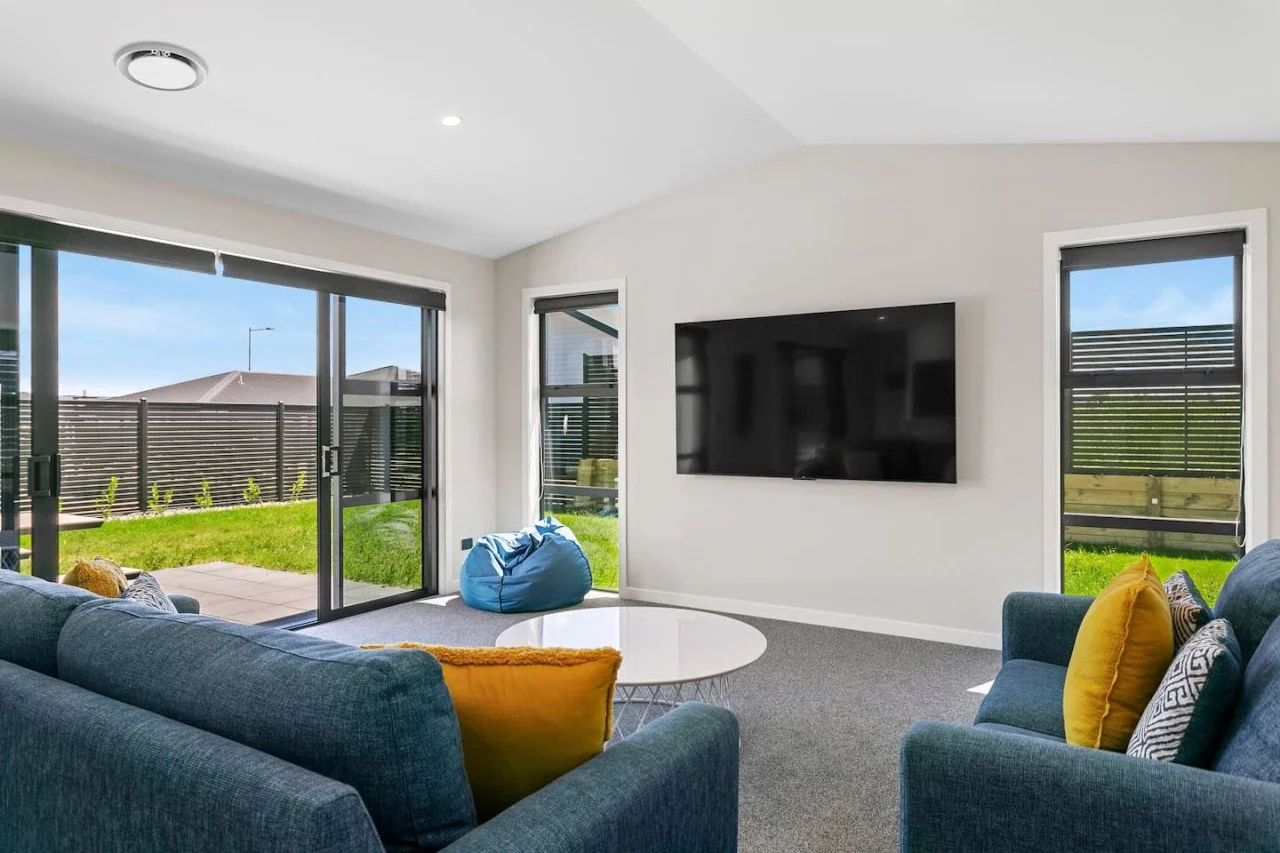 Bright living room with gray couch and yellow cushions, flat-screen TV on white wall, large glass sliding door leading to backyard with grass and wooden fencing, ceiling light, and a blue bean bag near the window.