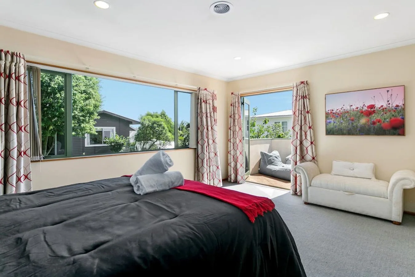A bedroom with large windows, a bed with black bedding and towels, a white sofa, and a sliding glass door leading to a balcony with outdoor cushions, decorated with curtains and a floral painting.