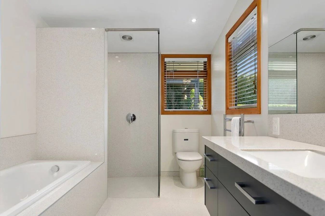 Modern bathroom with a bathtub, walk-in shower, toilet, double sink vanity, and two windows with wooden frames and blinds.
